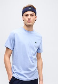 Νεαρός άνδρας που φορεί ανοιχτό γαλάζιο μπλουζάκι και σκούρα μπλε κορδέλα για το κεφάλι με πράσινο λογότυπο κροκόδειλου και στα δύο, στέκεται με τα χέρια στις τσέπες.