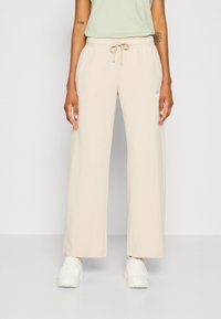 Beige wide-leg sweatpants with an elastic waistband and drawstring, featuring a small white logo on the left side. Soft fabric texture.