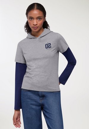 Felpa grigia con cappuccio e maniche corte, caratterizzata da una stampa "02" e un design a stelle, indossata sopra una manica lunga blu, abbinata a jeans blu.