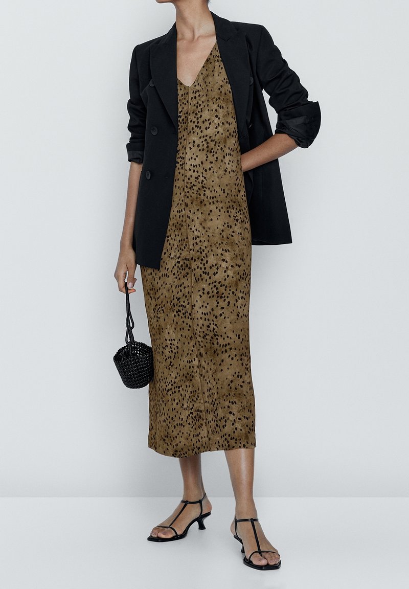 Blazer noir croisé sur une robe nuisette marron à pois, associé à des escarpins noirs à lanières et à un sac à main noir texturé.