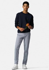Marineblaues Sweatshirt mit kleinem Logo, hellgraue Hose und weiße Sneakers. Das Model hält eine orangefarbene Blume und steht vor einem neutralen Hintergrund.