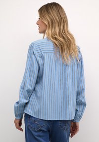 Camicia a maniche lunghe a strisce blu con linee verticali bianche e dorate, con polsini abbottonati e orlo arrotondato. Indossata sopra un paio di jeans in denim blu.