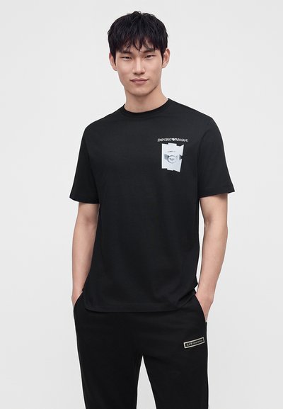 Άνδρας που φοράει μαύρο μπλουζάκι Emporio Armani με λευκό γραφικό και μαύρο παντελόνι με το λογότυπο EA7, στέκεται με τα χέρια στις τσέπες.