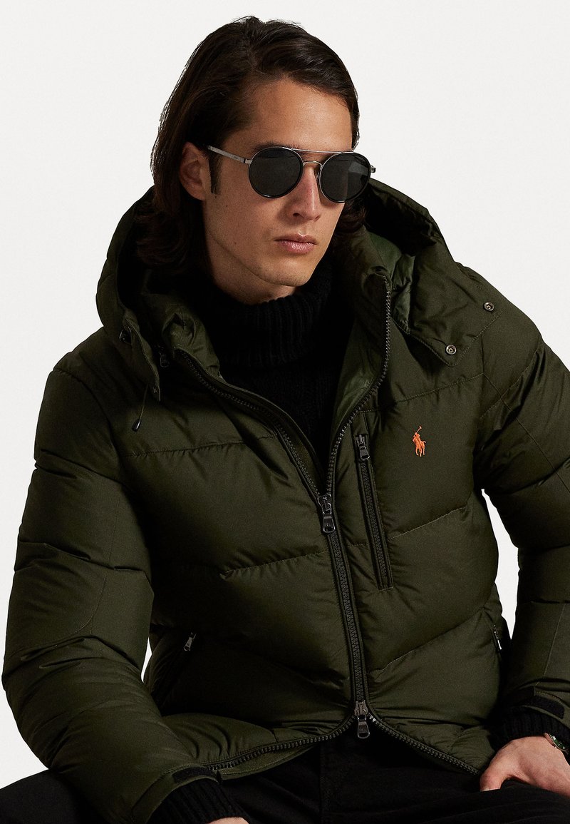 Groene gewatteerde jas met een capuchon, voorzien van een rits aan de voorkant en zijzakken. Draagt over een zwarte coltrui. Donkere zonnebril maakt de look compleet.