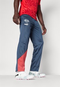 Blauwe sportbroeken met een rood zijpaneel, voorzien van een logo op de dij. Gepaard met witte voetbalschoenen met roze accenten en zwarte branding.
