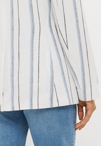 White shirt with blue and brown vertical stripes, made of textured fabric. Features long sleeves and a relaxed fit, partially tucked into blue jeans.