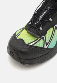 Sapatilha atlética preta e verde com um upper de malha texturizada, detalhes refletivos e sola flexível em borracha preta; apresenta um design com atacadores.