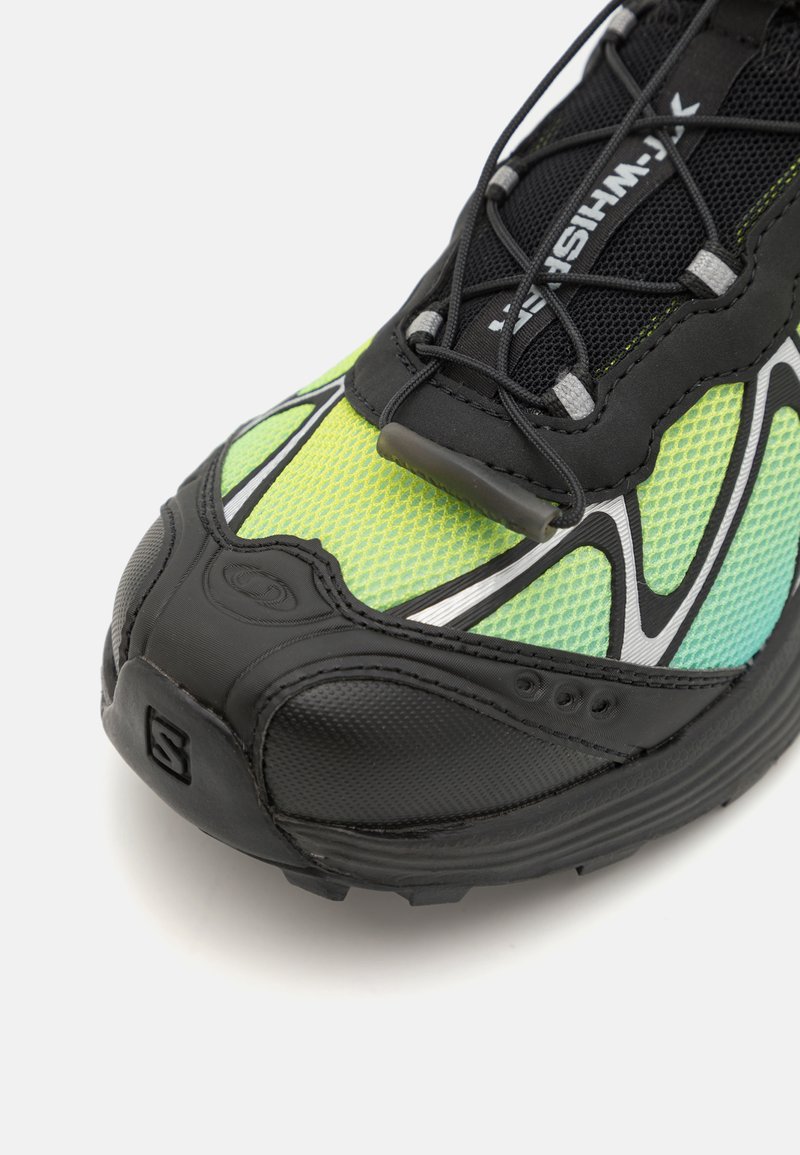 Zapatilla atlética negra y verde con parte superior de malla texturizada, acentos reflectantes y suela flexible de goma negra; cuenta con un diseño de cordones.
