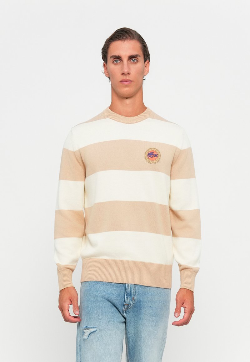 Beige och krämrandig tröja med rund hals och ribbade muddar, med en märkeslapp på bröstet. Buren med ljusblå jeans.