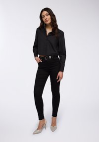 Maglietta nera cropped con chiusura a bottoni, pantaloni neri a vita alta e décolleté a punta di colore chiaro, abbinati a una collana in catena argentata.