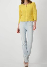 Gul stickad kofta med trekvartslånga ärmar och tre knappar, matchad med ljusblå jeans med broderade blommor vid manschetterna.