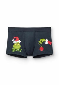 Boxer ajustados oscuros con la cara del Grinch llevando un sombrero de Santa a la izquierda y su mano verde sosteniendo un adorno rojo a la derecha.