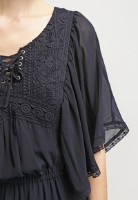 Mörkblå klänning med ett texturerat spetspanel, V-ringning, dekorativ snörningsdetalj och breda ärmar med spetskanter. Mjukt tyg faller lätt.