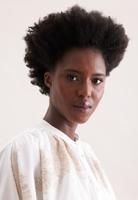 Blusa bianca con ricami floreali dorati, caratterizzata da un colletto alto e chiusura a bottone. Tessuto morbido con un design delle maniche leggermente a sbuffo.