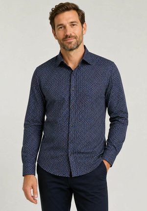 Man die een donkerblauw overhemd met lange mouwen draagt met een klein cirkelpatroon en een donkere broek, staand met één hand in de zak.