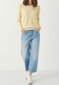 Femme portant un jean large bleu clair, un pull à manches longues rayé jaune et blanc, et des baskets blanches, debout les mains dans les poches.