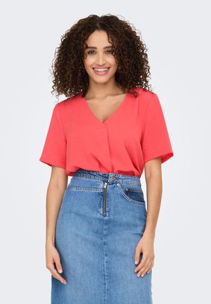 Glimlachende vrouw met krullend haar, gekleed in een koraalroze blouse met korte mouwen en V-hals, ingestopt in een blauwe denim rok met hoge taille en een rits aan de voorkant.