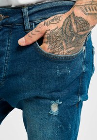 Jeans de mezclilla azul con corte slim, que incluyen un bolsillo frontal, costuras visibles, áreas desgastadas y un agujero deshilachado cerca del muslo.