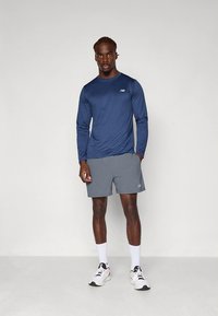 Tamsiai mėlyna ilgomis rankovėmis sportinė palaidinė derinama su pilkais šortais, pagaminta iš drėgmę sugeriančios medžiagos ir turi subtilų logotipą kairėje krūtinės pusėje. Balti kojinės ir sportbačiai.