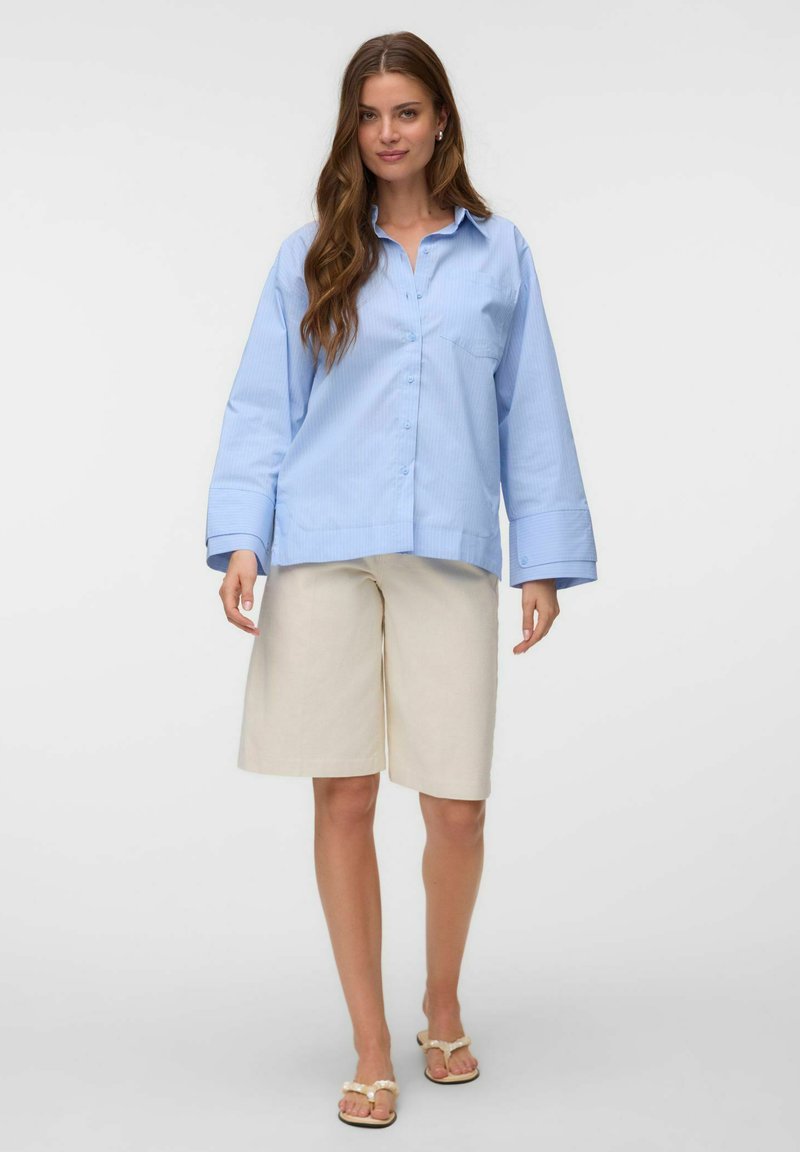 Femme aux longs cheveux bruns portant une chemise oversize bleu clair boutonnée, un short beige jusqu'aux genoux et des sandales beiges, debout sur fond blanc.