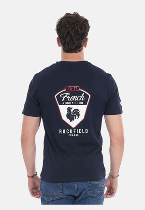 Homme portant un t-shirt bleu marine avec le logo du club de rugby français et un emblème de coq dans le dos, associé à un jean bleu, sur fond blanc.