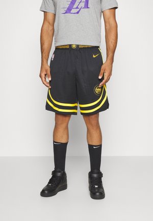 Mann trägt schwarz-gelbe Basketballshorts, graues LA-T-Shirt, schwarze Nike-Socken und schwarze High-Top-Sneaker und steht vor einer schlichten Wand.