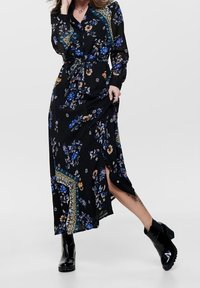 Robe maxi noire avec des motifs floraux en bleu et orange, manches longues, un design enveloppant, et une fente sur le côté, portée avec des bottines noires.
