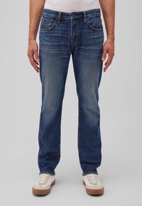 Hombre que lleva puesto unos clásicos pantalones vaqueros azul oscuro y zapatillas blancas, de pie contra un fondo gris liso.