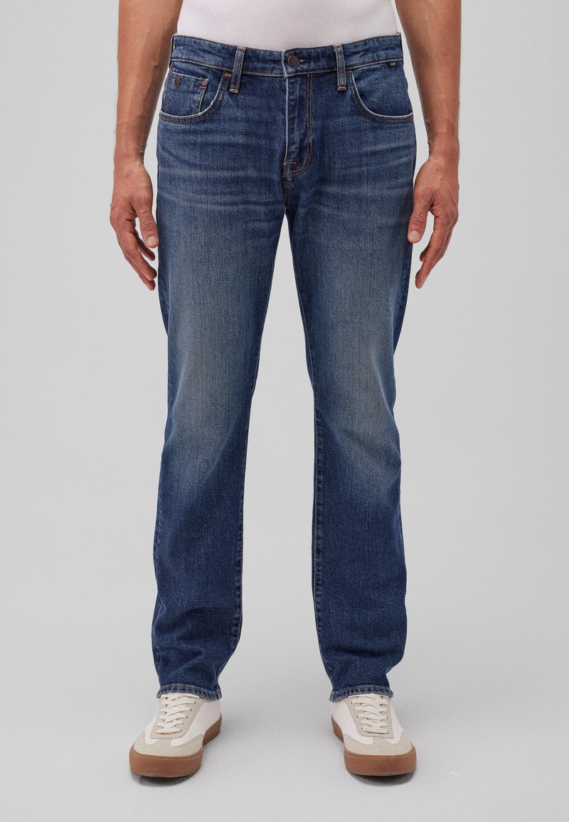 Hombre que lleva puesto unos clásicos pantalones vaqueros azul oscuro y zapatillas blancas, de pie contra un fondo gris liso.