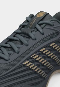 Σκούρο γκρι αθλητικό παπούτσι με χοντρές κορδόνια, πλεκτό ύφασμα και χρυσά ορθογώνια διακοσμητικά στη πλευρά κοντά στη σόλα.