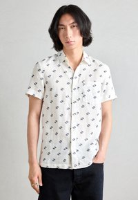 Short-sleeve, light-colored shirt made of fabric with a subtle texture, featuring a navy geometric pattern and a chest pocket.