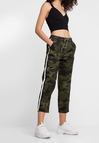 Camo broeken in donkergroen met witte zijkanten, met een trekkoord in de taille en een cropped fit. Gecombineerd met een zwarte cropped top en witte sneakers.