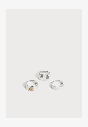 Trois bagues en argent sur fond blanc ; une bague possède un petit détail carré en or, une est simple, et une a une bande large et lisse.