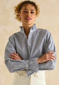 Sinine ja valge triibuline nööbitav särk, millel on volangkrae, pikad varrukad ja nööbiga mansetid, valmistatud siledast ja kergest kangast.