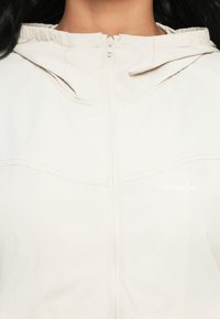 Gros plan d'une personne portant une veste zippée beige clair avec une capuche et un logo discret "Holzweiler" sur la poitrine.