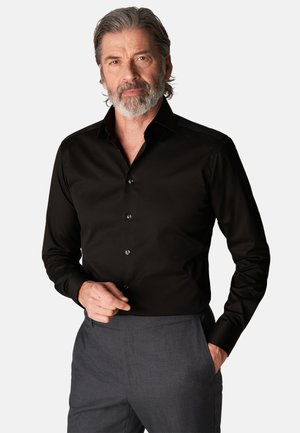 Svart skjorte med jevn tekstur, spiss krage og knappestolpe foran. Grå bukser er iført, med hendene plassert nær midjen.