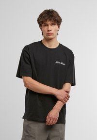 Black cotton t-shirt with short sleeves, featuring a small white "Kari Studios" text logo on the left chest area. Round neckline, relaxed fit.
