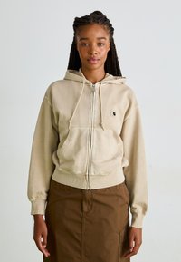 Béžová mikina na zips vyrobená z mäkkého materiálu, so prednou vreckom, kapucňou na sťahovanie a vyšitým logom na ľavej strane hrudníka.