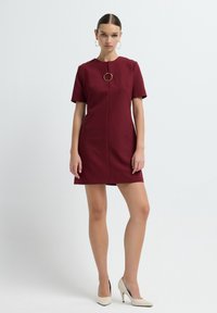 Femme debout dans une robe bourgogne à manches courtes avec une broche circulaire dorée au niveau du col et des chaussures à talons blancs sur un fond blanc.