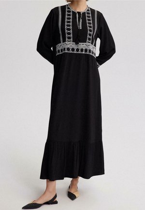 Robe longue noire à manches longues avec broderie géométrique blanche sur la poitrine et la taille, lien à pompons au niveau de l'encolure et ourlet volanté, portée avec des chaussures plates bicolores.