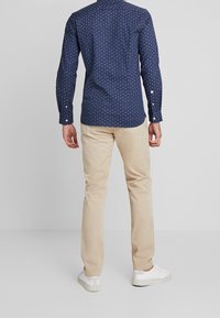 Tmavě modrá košile na knoflíky s malým bílým vzorem, doplněná béžovými chinos a bílými teniskami, pohled zezadu.