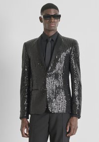 Blazer negro con lentejuelas, diseño entallado, solapas de satén y ajuste slim. Combinado con camisa y corbata negras, usado con gafas de sol.