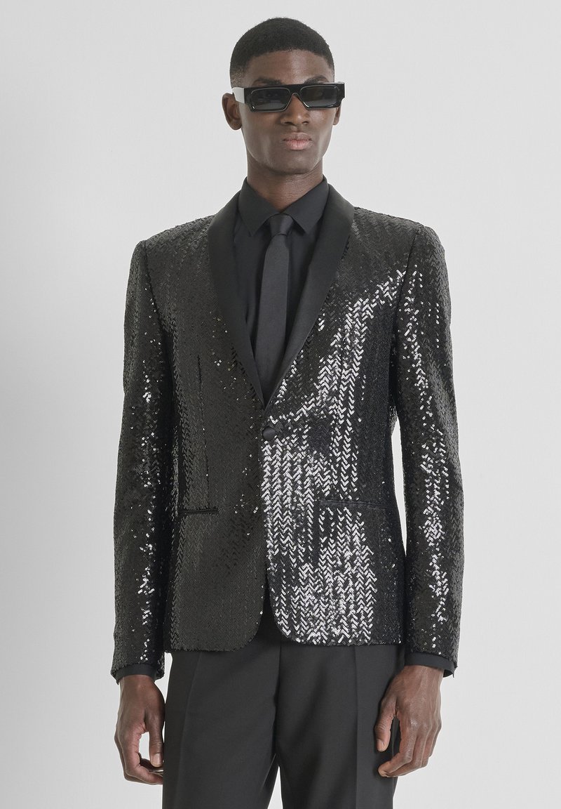 Blazer negro con lentejuelas, diseño entallado, solapas de satén y ajuste slim. Combinado con camisa y corbata negras, usado con gafas de sol.