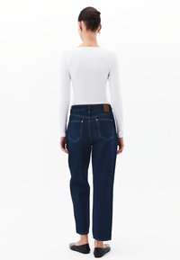 Jeans de tiro alto de color azul oscuro con un corte relajado, que presentan un parche de cuero marrón en la parte trasera y costuras sutiles en los bolsillos. Zapatos negros planos.