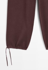 Pantalons en tissu marron foncé avec des poignets élastiqués et des cordons de serrage sur fond blanc.