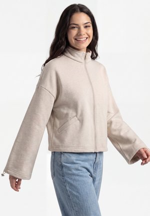 Ung kvinde smilende og iført en beige lynlåstrøje med brede ærmer og blå jeans mod en ensfarvet hvid baggrund.