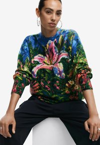 Maglione lavorato a maglia floreale colorato con vivaci gigli rosa, fogliame verde e uno sfondo blu. Vestibilità rilassata con polsini a costine.
