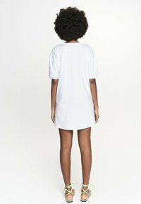 Biała bawełniana sukienka t-shirtowa z krótkimi rękawami, luźny krój i zaokrąglony dół. Noszona z zielonymi sandałami na paski, eksponującymi nogi. Widok z tyłu.