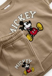 Beige sweatshirt och byxor set med en broderad Mickey Mouse grafik. Materialen är mjuka; sweatshirten har ribbade mudd, byxorna är elastiska.