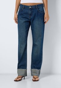 Jeans de ganga em azul escuro com corte reto. Apresenta bainhas dobradas com bordas desfiadas, cinco bolsos e um fecho clássico de botão.